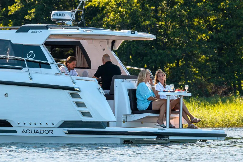 Two women enjoy drinks and snacks on a modern Aquador boat while two men relax inside, surrounded by greenery.